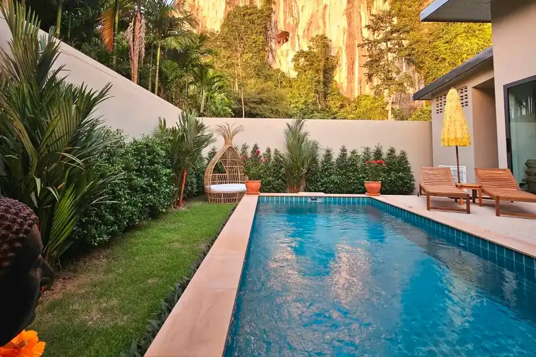 The swimming pool of a new 2 bedroom pool villa. an area of grass bordered by tropical shrubs and a high painted wall to the left. A patio with wooden sun lounger chairs and parasol in front of the house to the right. Beyond the far wall is a very high limestone cliff partly covered in vegetation bathed in the lat afternoon sun